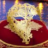 Masque vénitien intégral doré en métal finement découpé posé sur un coussin rouge avec broderies dorées