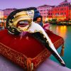 Masque vénitien long nez en plastique blanc et doré avec détails noirs et plumes rouges et violettes posé sur un coussin rouge brodé devant un canal de Venise au coucher du soleil