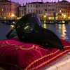 Masque vénitien médecin de la peste gothique noir en cuir avec rivets métalliques posé sur un coussin rouge brodé devant un canal de Venise au crépuscule